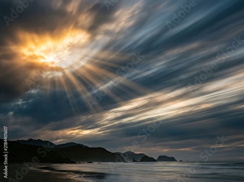 Sunbeams breaking through dramatic clouds over a misty coastline during sunset