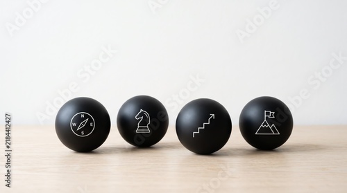 Four black spheres with white line icons representing business strategy, navigation compass, chess knight, growth stairs and mountain peak goal on wooden table surface