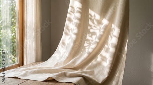 Beige linen curtain draped on wooden table near window with sunlight casting leaf shadows, minimalist interior design, natural fabric texture, cozy home atmosphere concept