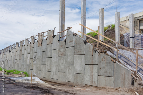 Concrete pillars rise from an active bridge construction site with plastic sheeting over earthworks, exposed support columns and nearby overpass, conveying industrial progress and structural repair wo