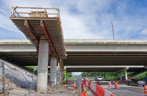 Bridge construction site on a highway overpass featuring scaffolding, concrete pillars, orange traffic barriers and cones, urban infrastructure work and road maintenance under a blue sky with clouds.
