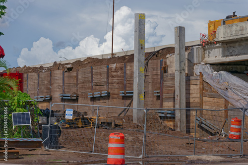 Concrete pillars rise from an active bridge construction site with plastic sheeting over earthworks, exposed support columns and nearby overpass, conveying industrial progress and structural repair wo