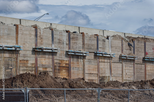 A weathered concrete retaining wall with steel braces towers above excavated dirt piles and temporary fencing at an urban construction site beneath a cloudy sky, conveying heavy infrastructure and ind