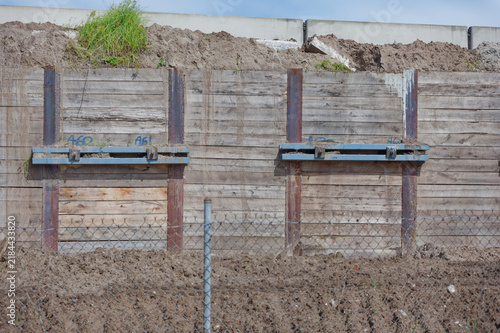 A weathered concrete retaining wall with steel braces towers above excavated dirt piles and temporary fencing at an urban construction site beneath a cloudy sky, conveying heavy infrastructure and ind