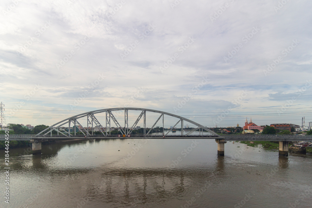 Obraz premium A steel arch bridge and elevated roadway cross an urban river beneath a broad cloudy sky with power lines and city buildings in the background.