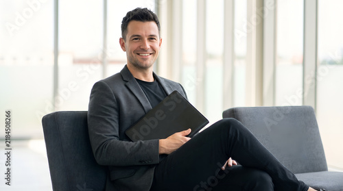 Smiling businessman holding closed laptop while sitting in office, entrepreneur, manager, executive, professional, workplace