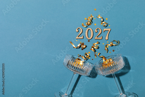 Champagne toast for 2027 New Year celebration with coupe glass, festive golden confetti stars and holiday decorations on blue background, top view.