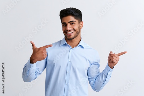 Young man in a light blue shirt pointing fingers to both sides against a plain white background