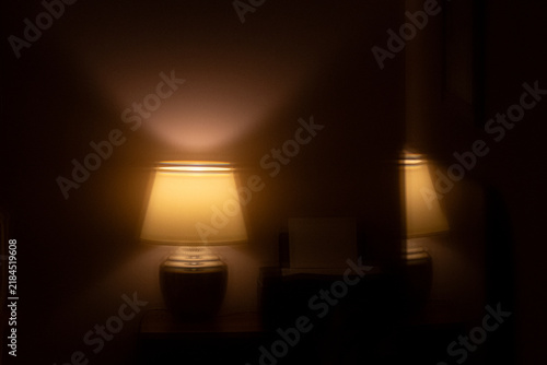 Warm illuminated table lamp casting soft amber light on a dark wall with a subtle mirror reflection. For introspection, mental health therapy, late-night insomnia, self-reflection, duality, and hope