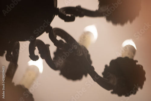 Silhouette of an antique wrought iron chandelier with burning candle-shaped lights in a warm, hazy, dark interior. Atmospheric low-key lighting creates a mood of mystery, vintage luxury, gothic