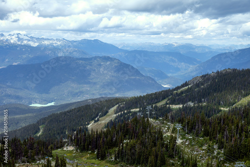 Expansive mountain vista with a ski lift traversing a forested slope under cloudy skies, Peak to Peak Gondola Whistler, Alder Creek, BC, Canada