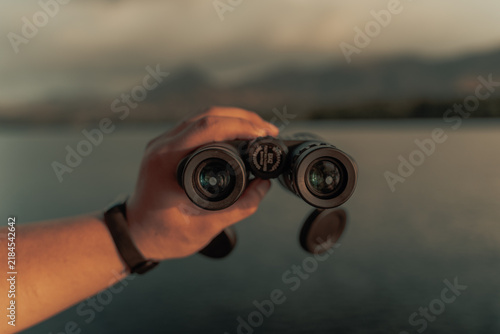 Konstfotografi Hand Holding Binoculars Overlooking a Scenic Mountain Lake at Sunset