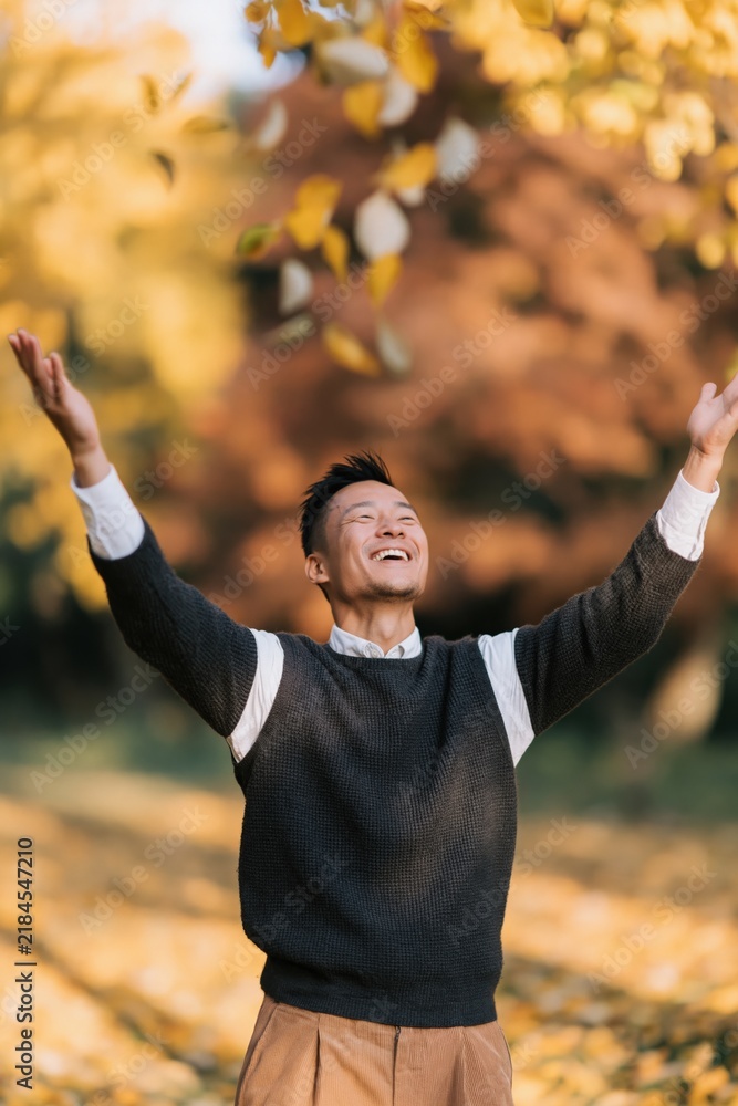 Fototapeta premium Cheerful man with arms raised amid swirling autumn leaves for fall banner and seasonal background featuring warm golden light and nature