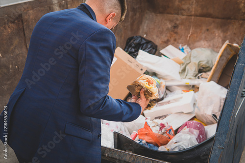 an impoverished businessman rummages in a trash can in search of food. A homeless homeless man in a suit holds burger in his hand while standing in a landfill. bankruptcy, no money concept