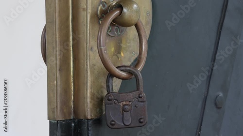 Old brass door with antique ring and padlock. Weathered metal surfaces show time and use. Dark door contrasts with golden hardware. Lock hangs securely on vintage door handle