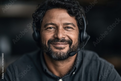 Smiling bearded man wearing a headset with microphone - close-up portrait suggesting a customer support or remote professional in a softly blurred workspace.
