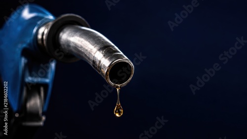 close-up of a blue fuel pump nozzle with a drop of gasoline dripping from its tip against a dark background.