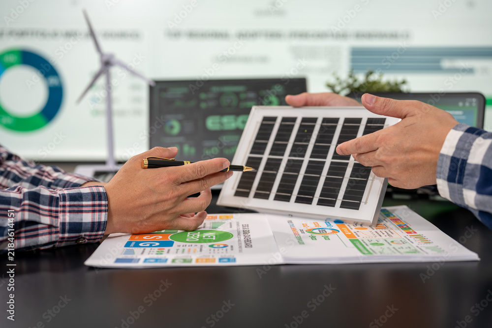Obraz premium Two people discuss a small solar panel over renewable energy charts and a laptop, with a wind turbine graphic in the background.