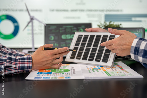 Two people discuss a small solar panel over renewable energy charts and a laptop, with a wind turbine graphic in the background.