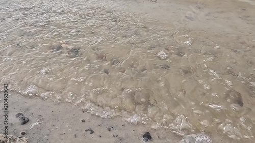 A gentle wave washes over a sandy beach scattered with small pebbles and shells, foam spreading forward then receding steadily across the wet shore under natural daylight.