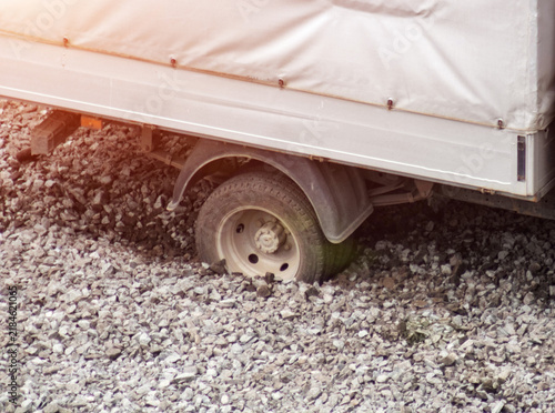Foto Trailer wheel deeply stuck in loose gravel of unpaved road, vehicle experiencing traction loss and immobility, sunlight effect highlighting scene of transport challenge