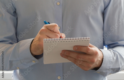 Close view of an unrecognizable man in a light blue shirt holding a pen and writing in a spiral notepad, suggesting note taking, reporting, or business work
