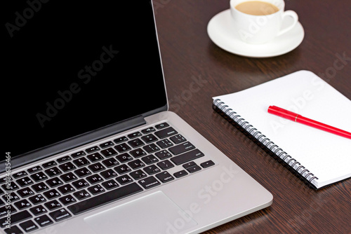 Close view of a laptop with a blank black screen beside a spiral notebook, red pen, and coffee cup on a dark wooden desk in a simple workspace setting