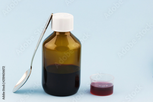 Amber glass medicine bottle partly filled with red liquid beside a clear measuring cup and metal spoon on a light blue studio background