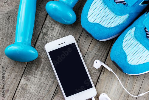 Top view of blue running shoes, a smartphone with blank screen, earphones and a dumbbell arranged on a weathered wooden floor for fitness and workout concepts
