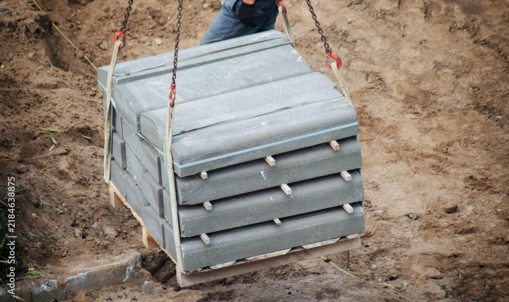 Fototapeta premium Concrete curb stones on pallet lifted by manipulator chains for construction work. Road parts transported over brown earth. Infrastructure development material movement unloading process.