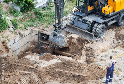 Excavator bucket digs earth trench for foundation work. Construction machinery moving soil during utility installation or building site preparation. Worker observes digging process 