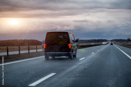 Rear view of black passenger van moving on multilane highway during evening hours, vehicle driving on wet asphalt road surface under cloudy sky, road transportation concept, evening sunset. 