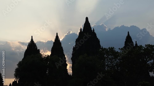 Silhouetted temple peaks reach toward a radiant sky. Sunlight spills through clouds, casting sacred rays. Ancient architecture stands still against a quiet horizon. Mystical glow suggests reverence