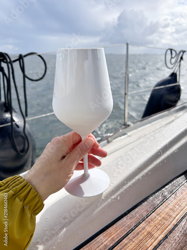 A hand holds an opaque white wine glass aboard a sailing boat with open sea and cloudy sky in the background