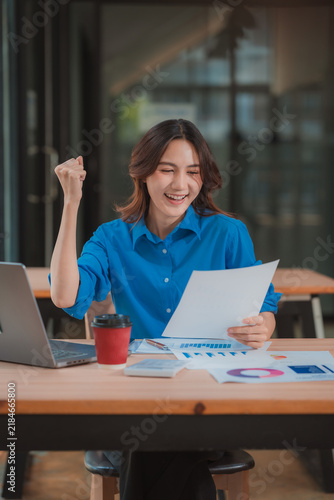 Excited Asian businesswoman raising arm celebrating business success while holding document at office desk