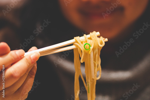 Close-up of a happy woman using chopsticks to eat delicious ramen noodles with a smiling face in the background.
