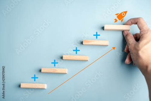 Business growth startup launch and success strategy. Hand placing wood block step on stairs with rocket icon and upward arrow.