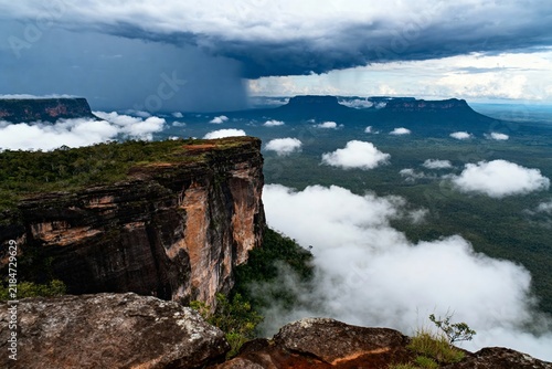 A dramatic cliff edge overlooks a sea of clouds with distant plateaus under stormy skies.