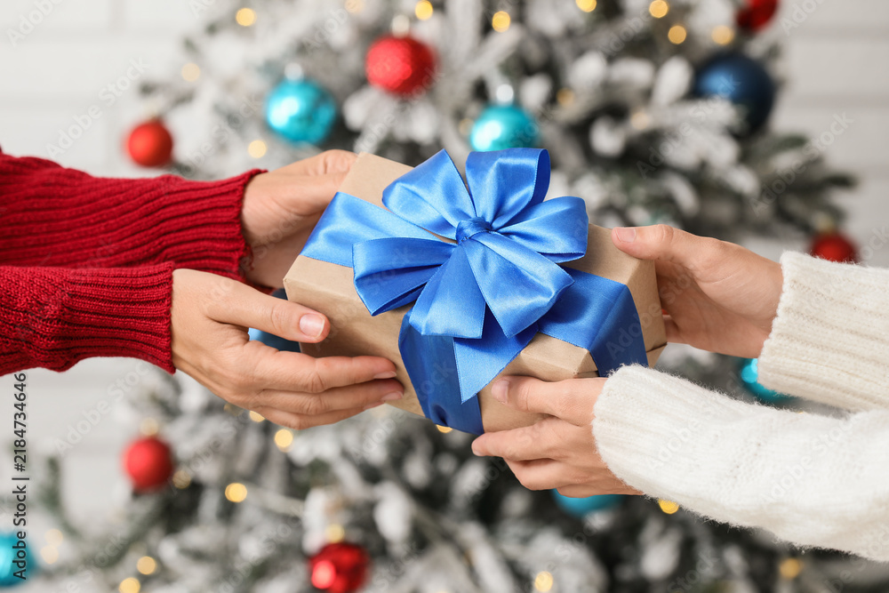 Fototapeta premium Holiday gift exchange, people giving a present with blue ribbon in front of a decorated Christmas tree for seasonal celebration and winter festivities
