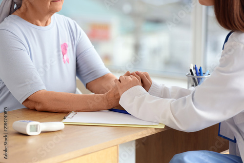 Senior woman with pink ribbon and doctor holding hands in clinic. Breast canc...