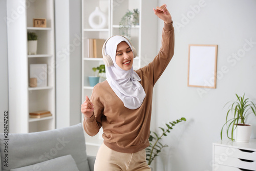 Beautiful young Muslim woman in hijab and headphones in living room