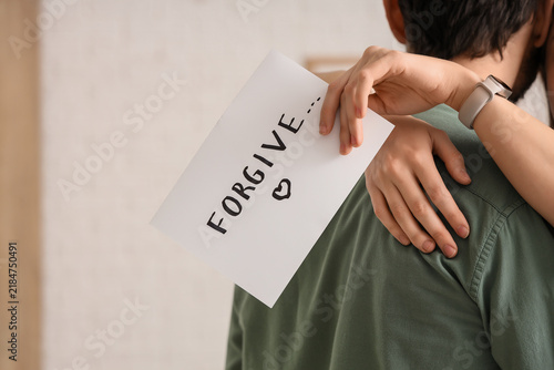 Young woman holding paper with word FORGIVE and hugging her husband at home, ...