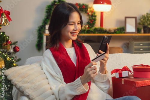 Cute Asian young woman wearing a white knitted sweater and red scarf, sitting on a sofa in the living room while using a smartphone and credit card for online shopping in Christmas day.