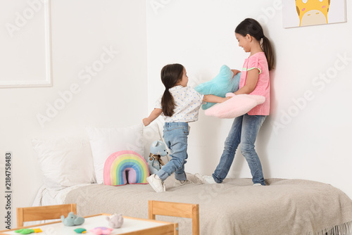 Cute little Asian happy sisters fighting pillows on bed at child room