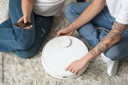 Young couple sitting on floor with modern robot vacuum cleaner and mobile pho...