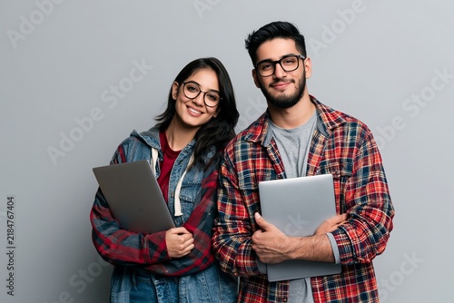 Young man and woman holding laptops against a grey studio background wearing casual checkered shirts