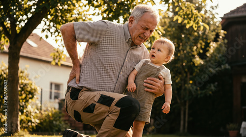 Grandfather holding toddler outdoors while experiencing back pain and maintaining caring family bond