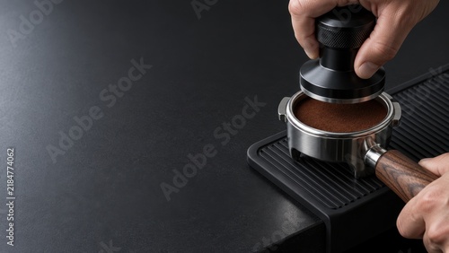 Close up of hand tamping ground coffee in portafilter with tamper on black surface beside espresso machine