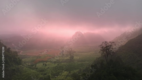Atmospheric view of a lush mountain valley covered in soft pink morning mist, with layered hills, forested slopes, and a tranquil natural landscape.