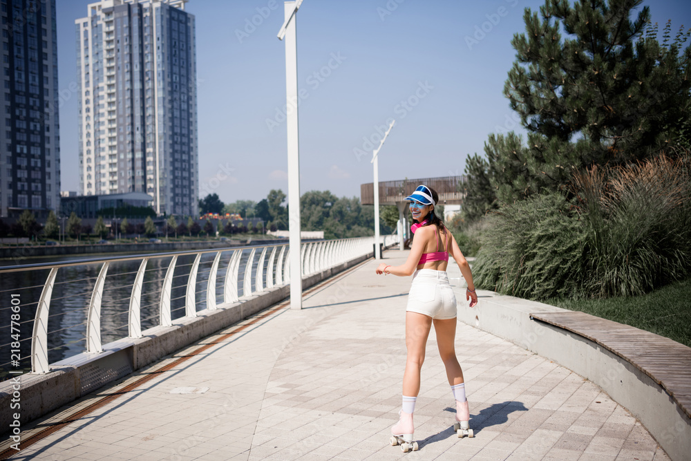 Fototapeta premium Young woman roller skating along an urban waterfront promenade in summer wearing a visor and headphones, smiling back while enjoying outdoor fitness and retro sports style